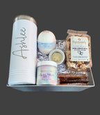 Birthday gift set with a monogrammed tumbler, bath bomb and scrub, and gourmet snacks on a wooden surface