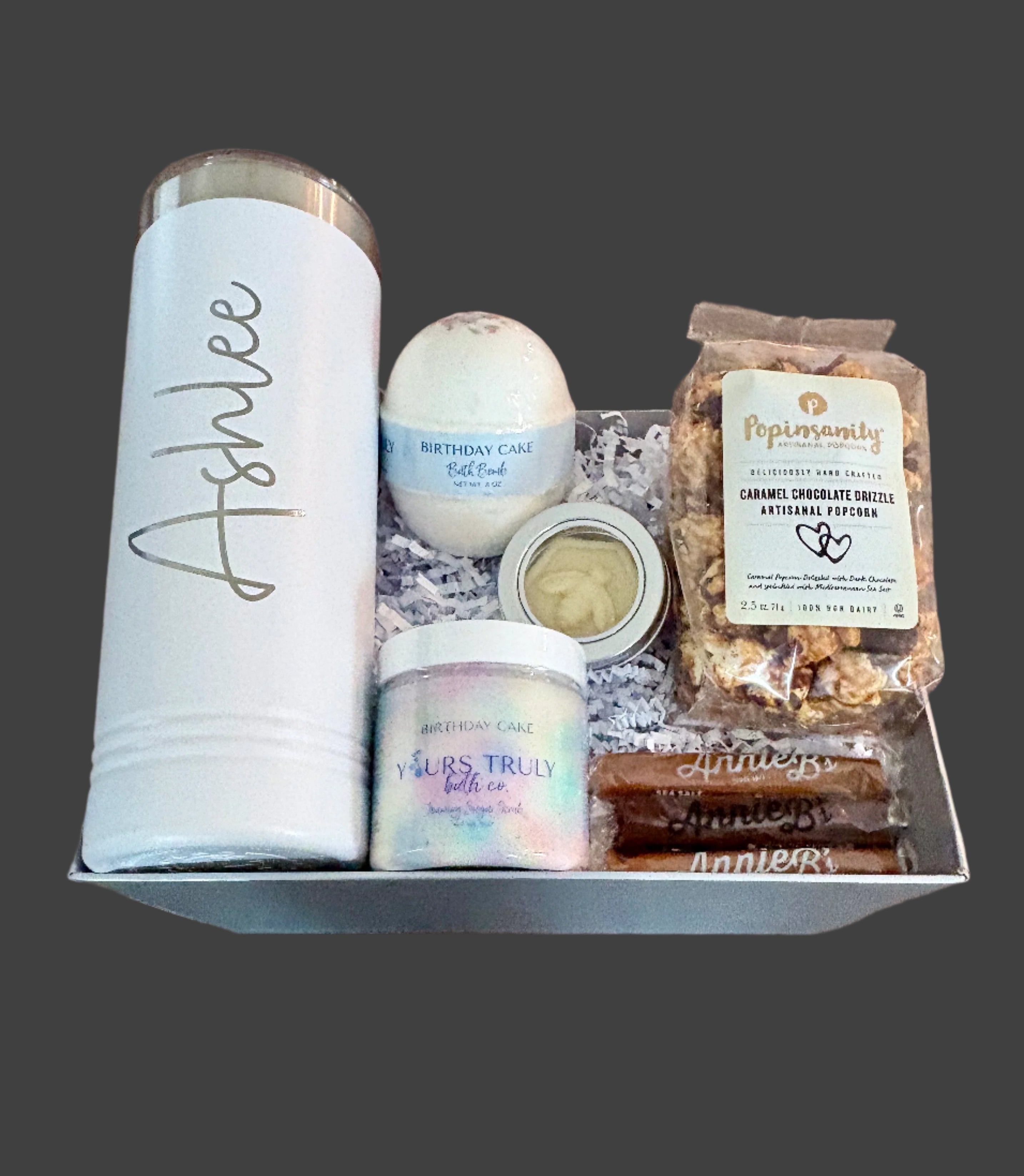 Birthday gift set with a monogrammed tumbler, bath bomb and scrub, and gourmet snacks on a wooden surface