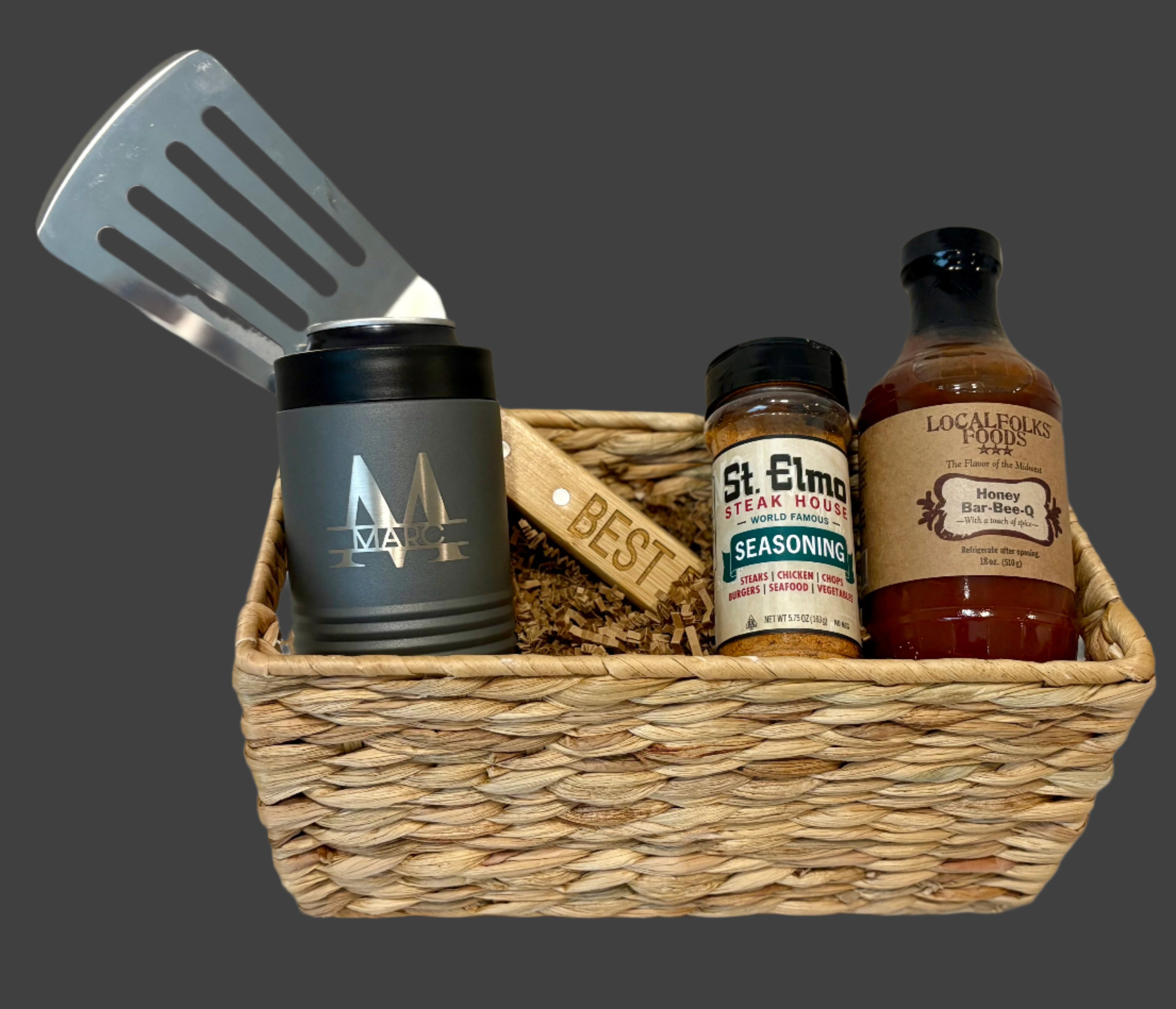 Basket with barbecue-themed items including a spatula, canister, seasoning jar, and bottles on a neutral background.